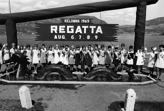 A line of women posed with their right hands held up, standing behind the statue of a sea serpent. They wear dresses, sashes and crowns. There is a sign above them. Text: Kelowna 1969 Regatta Aug. 6.7.8.9