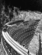 A train travels across railway tracks suspended by wooden beams on steeply inclined ground. A person stands on the tracks in the bottom right hand corner.