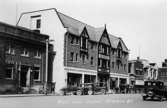 A brick building with cars parked in front of it as seen from the road. Other buildings line the street alongside the building. Text: Royal Anne Hotel Kelowna BC