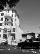 A tall building under construction next to a much smaller building as seen from the street. There is a modern truck parked on the road.