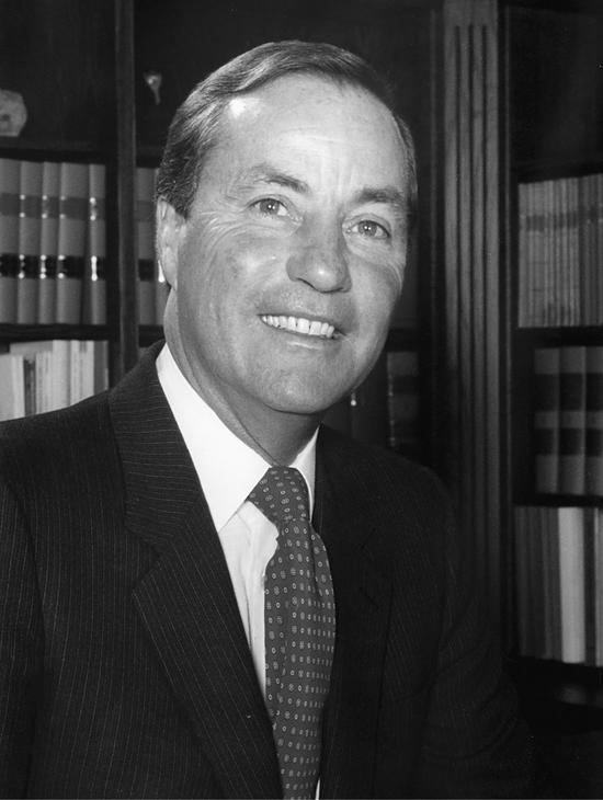 A portrait of a man with his body facing right but head turned to the camera. His gaze lands above the camera. He is in a dark suit, a collared shirt and a tie. Behind him are shelves of books.
