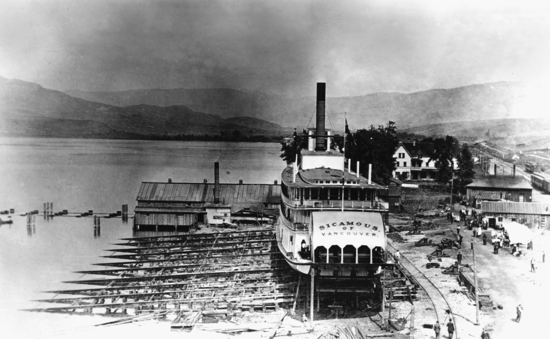 A large boat docked on land and suspended. Below, people are working and behind them are buildings along a road.