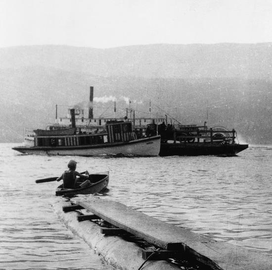 A person paddling in a small boat watching a steamboat push a floating structure carrying an old car.