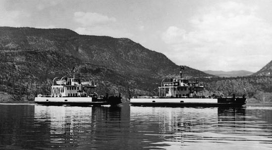 Two ferries on a body of water. In the background are tree-covered hills.