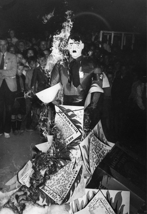 A figure of Adolf Hitler on fire. On him are sheets of paper with images and text. Behind the figure is a crowd watching, including children. Text: Help Burn Hitler. Add a Flame.