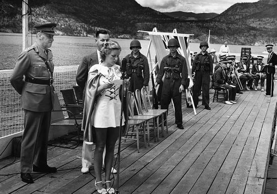 A light-haired woman in a short dress, heels, a sash and a cape reads from a piece of paper in front of a microphone. Behind her, a band and men in military uniforms watch. They are on a dock. 