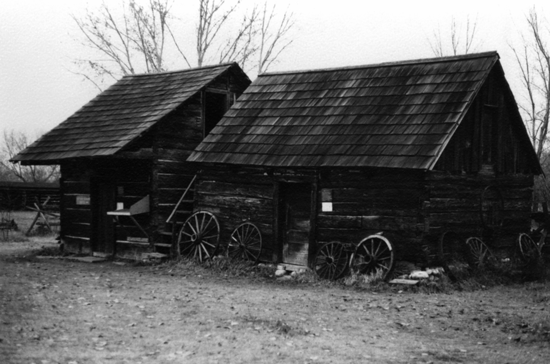 Two older wooden buildings. Several cart wheels lean against one of the buildings. In the background are trees with a few leaves.