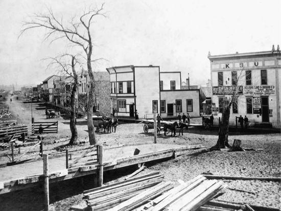 A pile of wooden planks lie on the ground in front of a bridge. Behind them are horse-drawn carts on a dirt road and light-coloured buildings.
