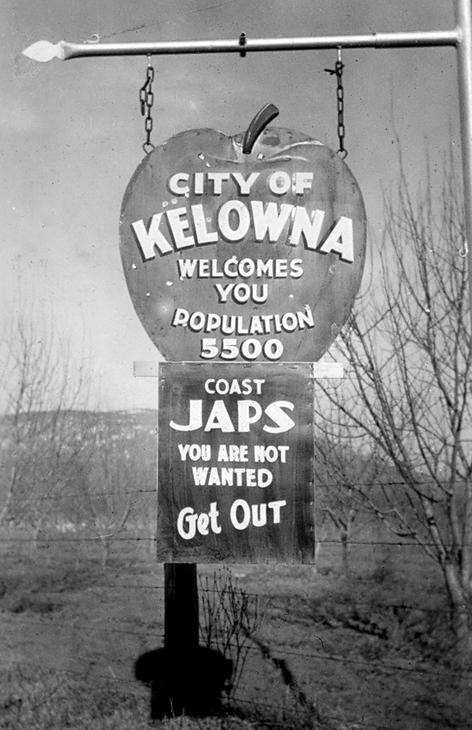A sign hanging from a metal pole. The top of the sign is in the shape of an apple and the bottom, a rectangle. Text: City of Kelowna Welcomes You Population 5500, Coast Japs You Are Not Wanted Get Out