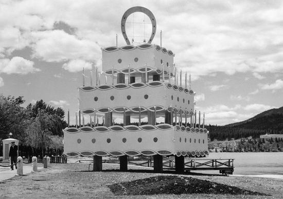A large statue of a three tiered cake on grass with candles circling each tier's perimeter. A large horseshoe sits at the top of the cake.