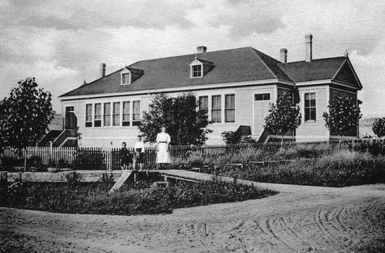 A tank parked on grass. Behind it is a wooden building with a Canadian flag in front of it. Text: Brigadier Angle Armoury