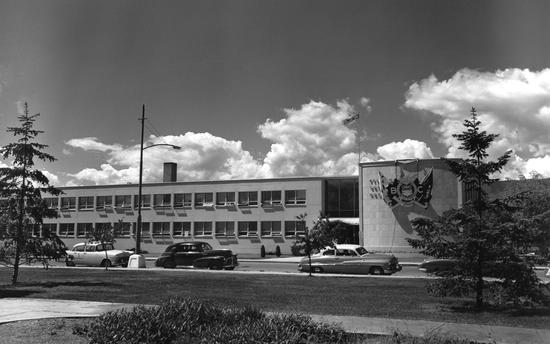 A long two storey building displaying a large crest and flags by its entrance, as seen from across the road. Old cars are parked on the street.