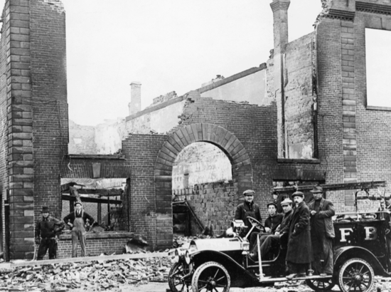 A damaged brick building with no roof. People stand in front of it, some in a old car.