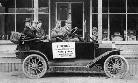 Five men sitting in an old car parked in front of a shop while looking at the camera. There is a sign on the car. Text: Simeon's Okanagan Mission Auto Stage, Single Fare 50 cents, Return 75 cents