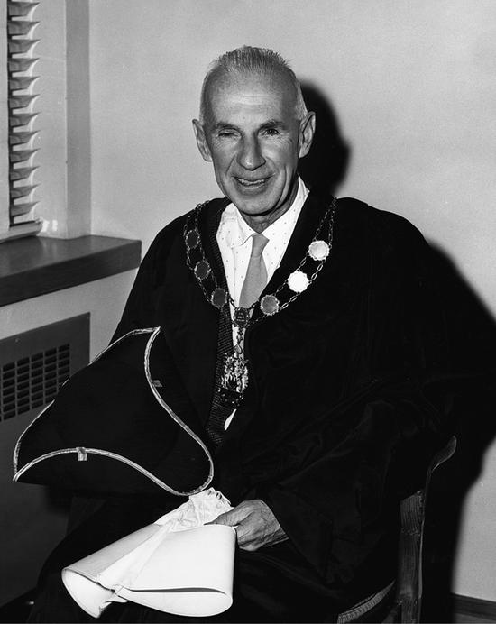 A man sits turned to the left while smiling at the camera. He wears a collared shirt, a tie, a large metal necklace and a cloak. In his hand, he holds a hat.