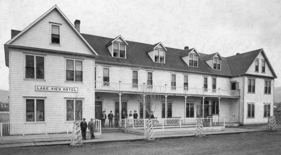 A large, light-coloured wooden building. Eight people stand in front of it looking at the camera. Text: Lake View Hotel