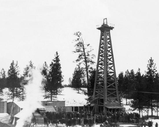 Numerous people standing before a tall metal structure. Beside the structure are wooden buildings. In the background are trees, and snow lines the ground.