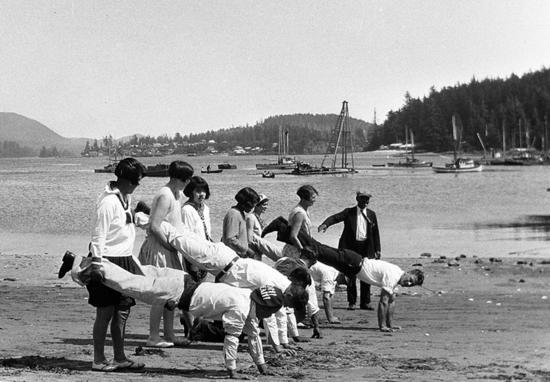 Wheelbarrow race at Clayoquot Days on Stubbs Island, early 1930s. People travelled from all over Clayoquot Sound to participate in this May 24 event, the largest, most important gathering of the year. Sporting events of all kinds, boat races, and a  community picnic drew hundreds of people: aboriginals, Japanese, and European settlers alike. The growing town of Tofino can be seen in the background, also Bill Bond’s pile driver.